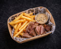 Basket of fries and sliced meat with a dipping sauce on a dark background